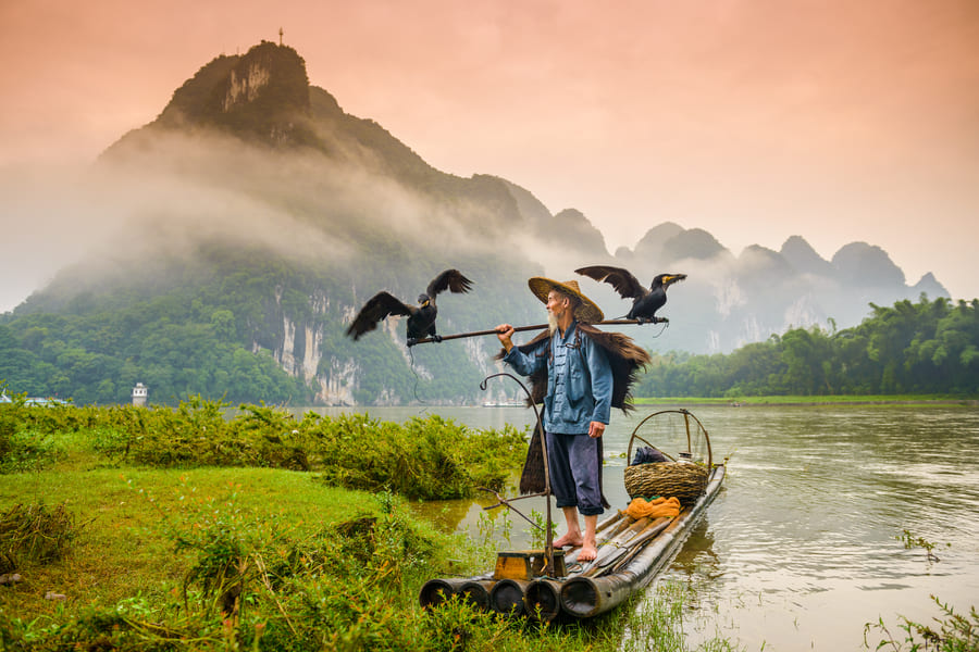 Pescador de cormonrán sobre una lancha en el río Yulong en Yangshuo con montañas de fondo